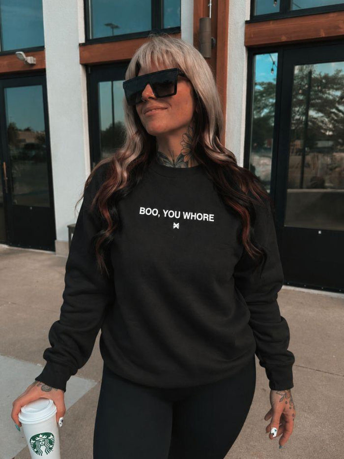A woman wearing a black crewneck sweatshirt with the white text 'BOO, YOU WHORE' and a small 'M' graphic below it, paired with black pants and sunglasses, holding a Starbucks coffee cup outdoors.