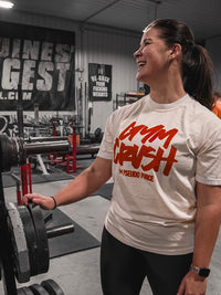 Beautiful woman standing in a gym wearing a cream-colored T-shirt. Front of the shirt features red handwritten-style text reading ‘Gym Crush’ with ‘Pseudo Force’ printed below.