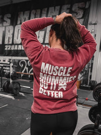Beautiful woman standing in a gym wearing a red crewneck sweatshirt. Back of the sweatshirt features large white text reading ‘Muscle Mommies Do It Better’ with a Pseudo Force symbol.