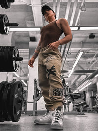 Person in a gym wearing a tank top and tan jogger sweat pants with visible pseudo force branding.