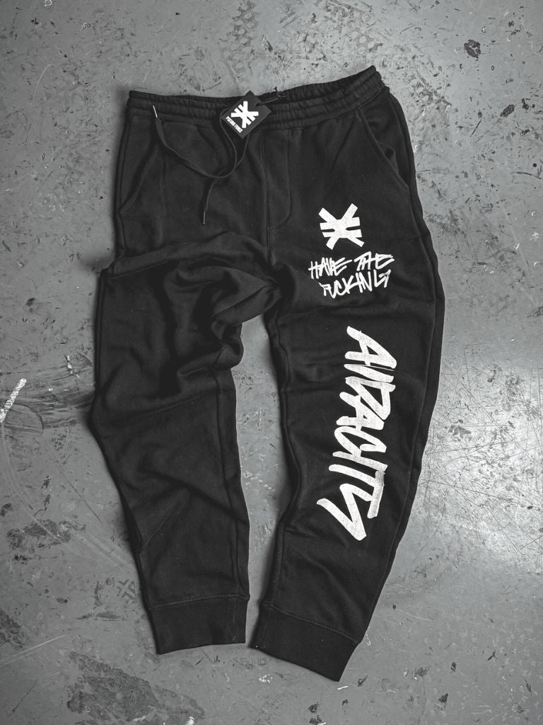 Black sweatpants laid flat on a concrete floor. White graphics include a symbol and the phrase ‘Have the fucking audacity’ on the upper leg, with the word ‘Audacity’ printed vertically down the lower leg.