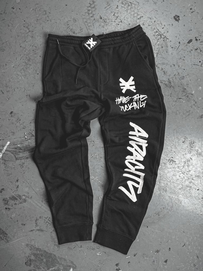 Black sweatpants laid flat on a concrete floor. White graphics include a symbol and the phrase ‘Have the fucking audacity’ on the upper leg, with the word ‘Audacity’ printed vertically down the lower leg.