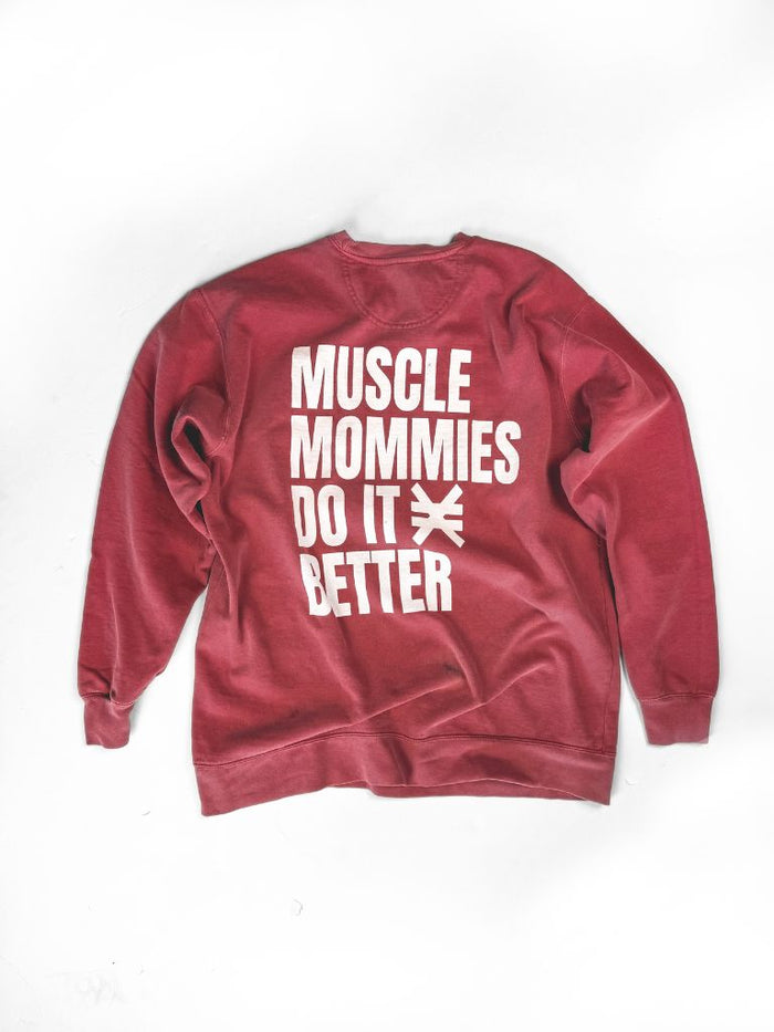 Red crewneck sweatshirt laid flat on a white background. Back of the sweatshirt features large white text reading ‘Muscle Mommies Do It Better’ with a Pseudo Force symbol between the words.