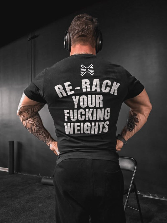 Person standing in a gym wearing a black T-shirt. Back of the shirt features white text reading ‘Re-rack your fucking weights’ with a Pseudo Force symbol above the text.