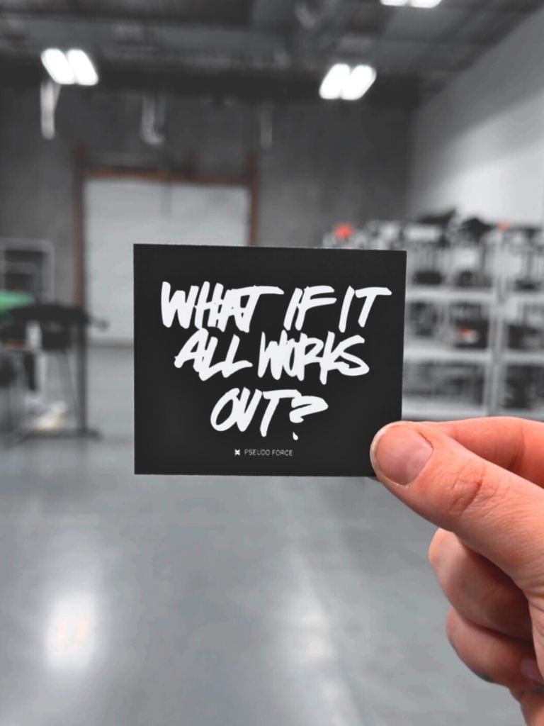 Hand holding a black square sticker that reads ‘What if it all works out?’ in white handwritten-style lettering. Small ‘Pseudo Force’ text appears below the phrase. Background shows a blurred warehouse interior.