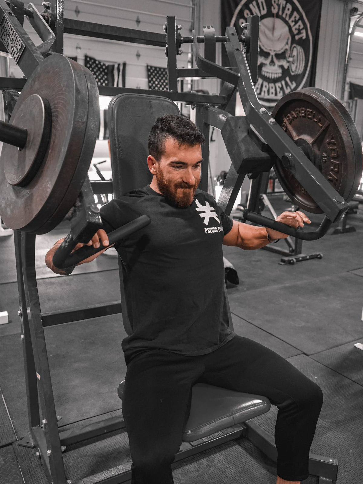 Man seated on a chest press machine in a gym wearing a black Pseudo Force T-shirt. Shirt features a small white Pseudo Force symbol and logo on the left chest.