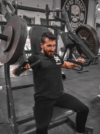 Man seated on a chest press machine in a gym wearing a black Pseudo Force T-shirt. Shirt features a small white Pseudo Force symbol and logo on the left chest.