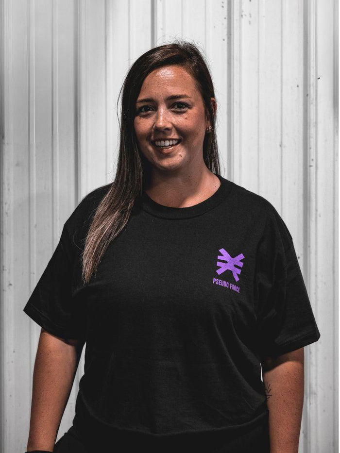 A smiling woman with long brown hair wearing a black t-shirt with a purple Pseudo Force logo on the left chest, standing against a white corrugated metal wall.