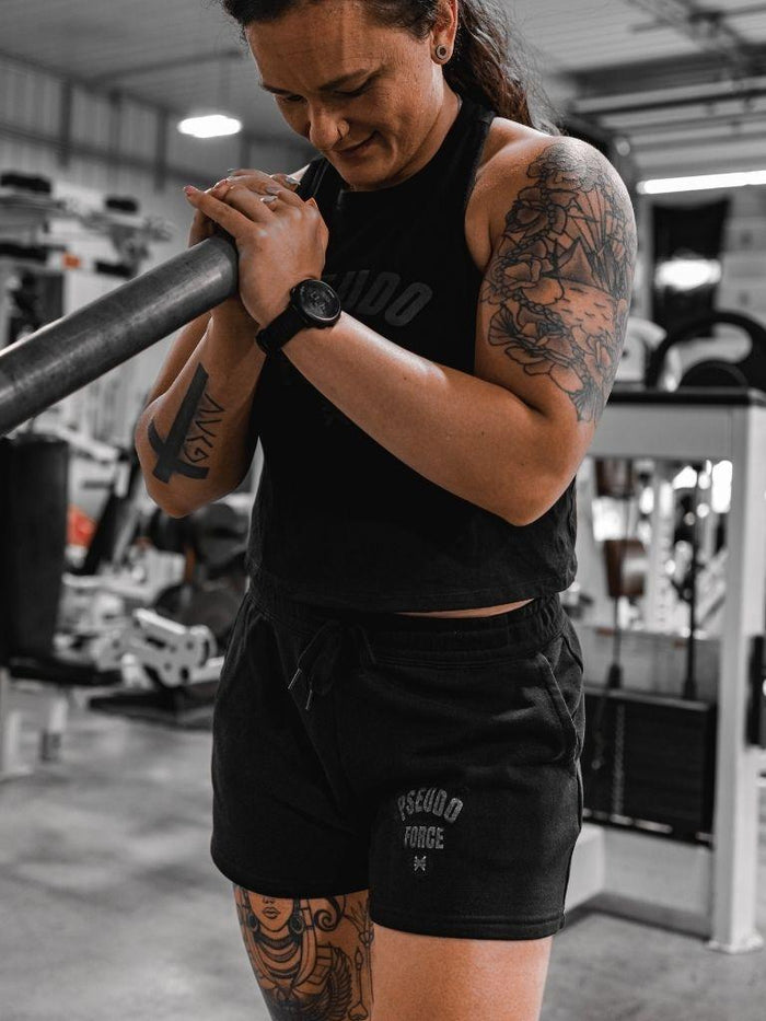 A woman in a gym wearing a black tank top and black shorts with white text, holding a barbell. The shorts say pseudo force with the logo under, in a lighter gray for a blackout effect.