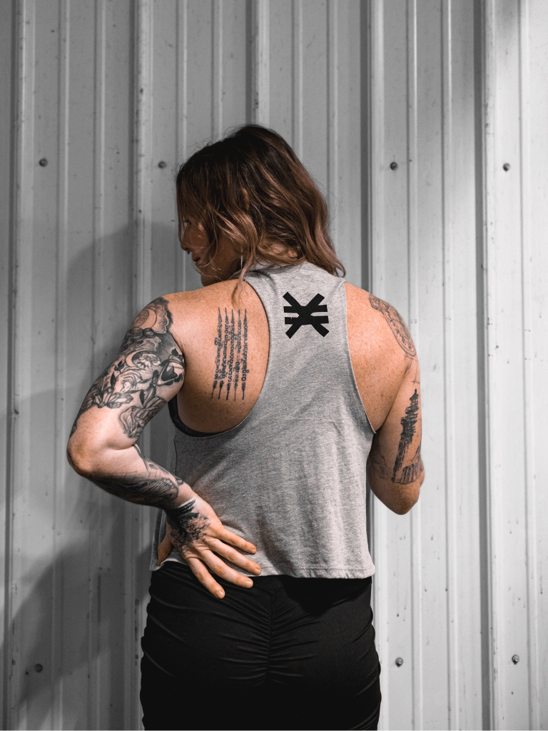 Person wearing a gray tank top with a logo, standing against a metallic wall.
