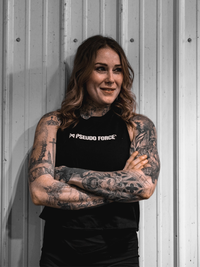 Person with tattoos wearing a black 'Pseudo Force' shirt against a gray wall.