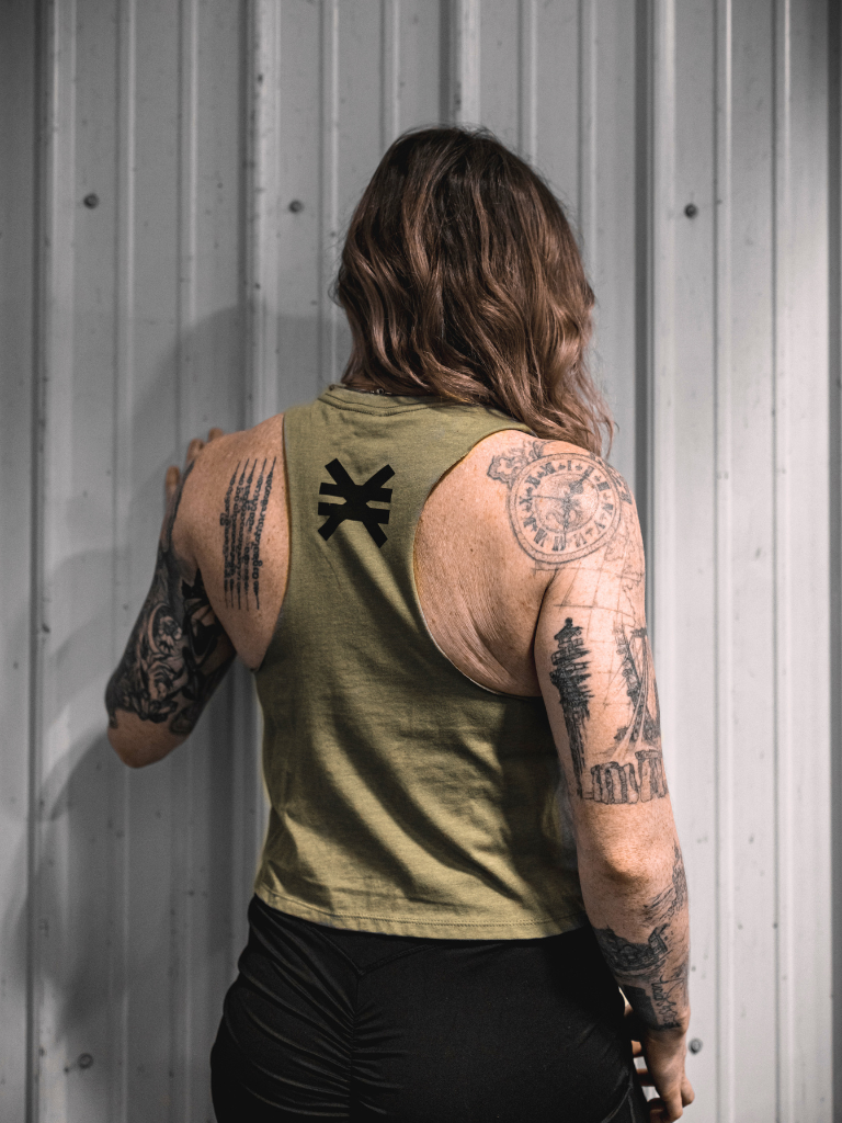 Person wearing a green sleeveless shirt with visible tattoos on arms against a metallic wall.