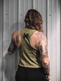 Person wearing a green sleeveless shirt with visible tattoos on arms against a metallic wall.
