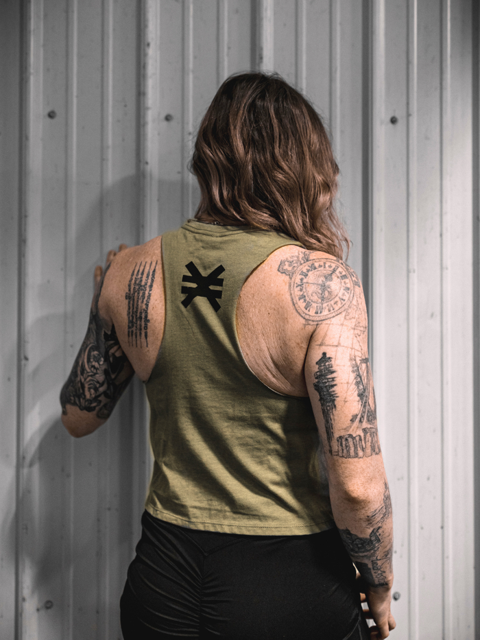 Person wearing a green sleeveless shirt with visible tattoos on arms against a metallic wall.