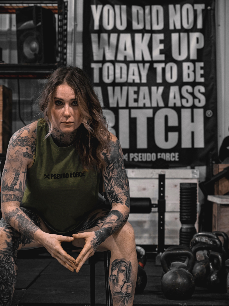 Woman with tattoos wearing a green tank top in a gym setting with motivational poster.