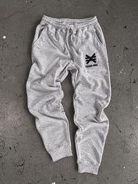 Gray jogger sweatpants with a black pseudo force logo on the left leg. Laying on cement floor with ink splatters.