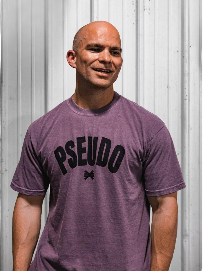 A man wearing a heathered purple t-shirt with the word 'PSEUDO' printed in black across the chest, standing against a light gray corrugated metal wall.