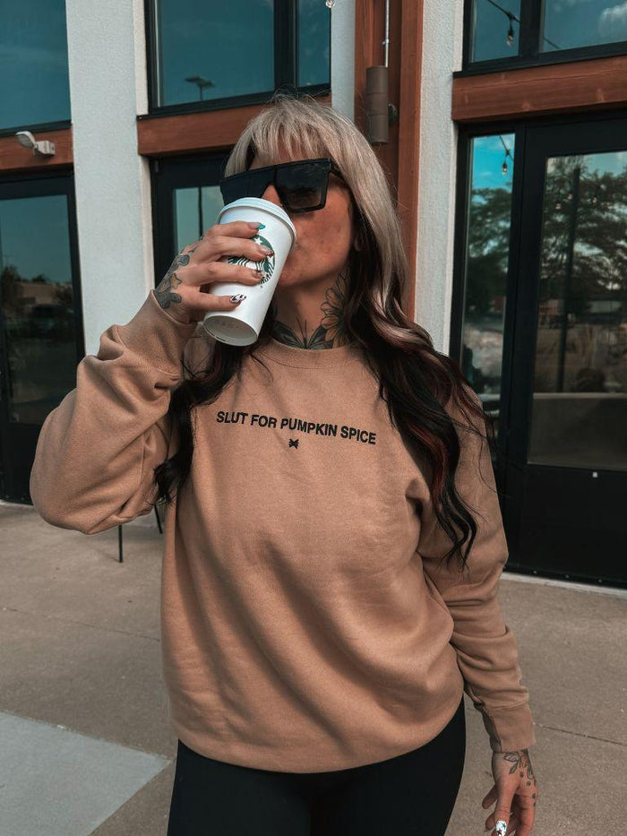 A woman wearing a tan crewneck sweatshirt with the text 'SLUT FOR PUMPKIN SPICE' printed in black across the chest, holding a white Starbucks coffee cup. She is wearing black sunglasses and black pants, standing outdoors in front of a modern building with glass doors.