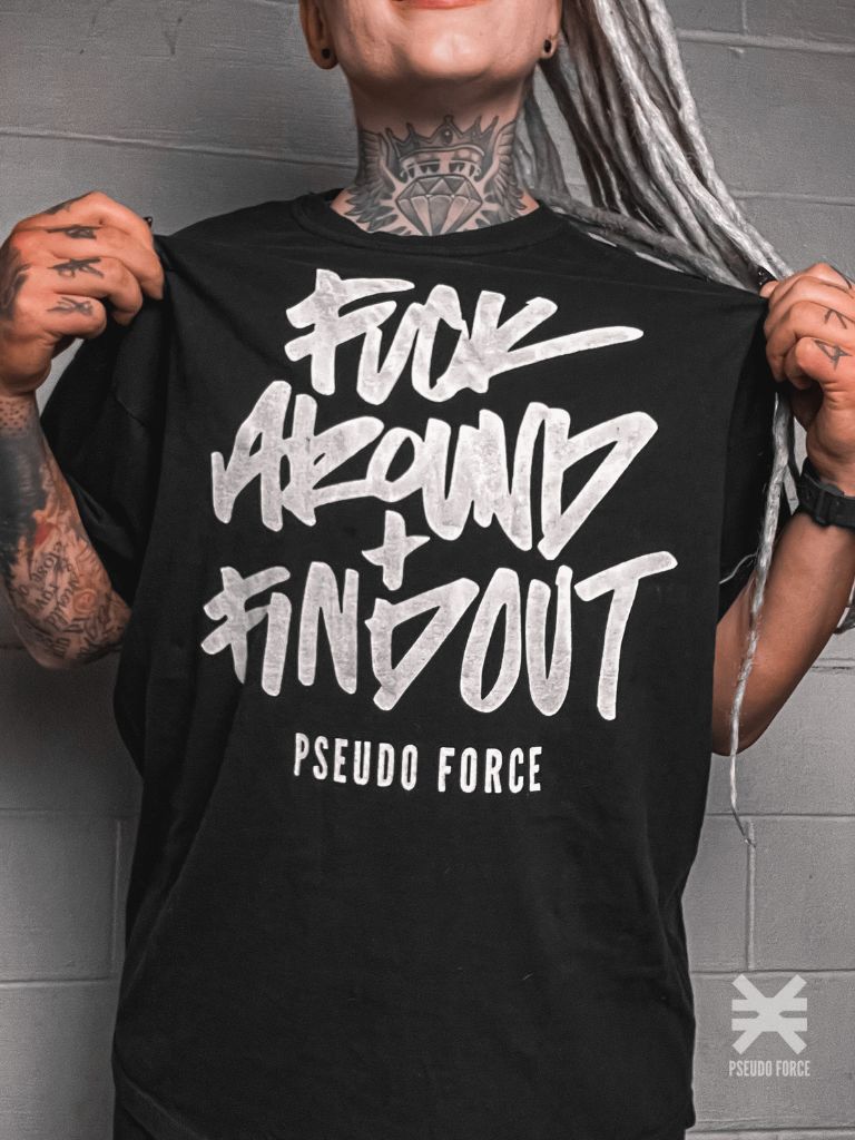 This image features a bold black t-shirt with the phrase "FUCK AROUND + FIND OUT" written in large, white, distressed lettering, followed by "PSUEDO FORCE" in a smaller font. The t-shirt is modeled by an individual with tattoos, highlighting the edgy and youthful appeal of the garment.